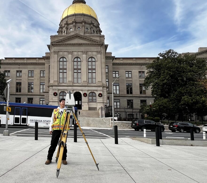 Surveyor standing behind a survey mapping camera on a city sidewalk. Behind them, a city bus passes and a large city building can be seen.